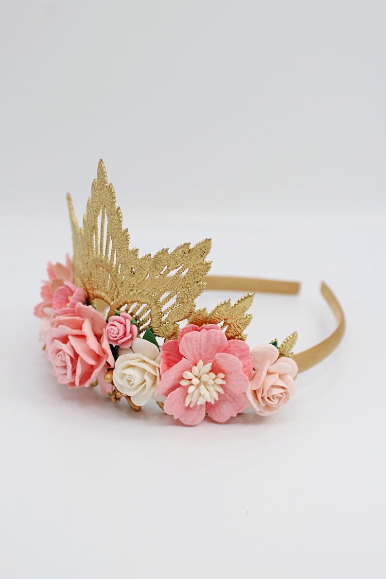 May include: A gold crown headband with pink and white paper flowers. The crown is made of gold lace and has a delicate design. The flowers are arranged around the crown and are accented with gold beads.