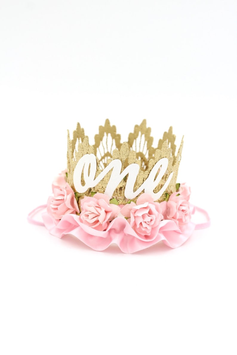 Puede incluir: Una corona de purpurina dorada con flores de tela rosa y la palabra "uno" en letras blancas. La corona est&aacute; dise&ntilde;ada para el primer cumplea&ntilde;os de un ni&ntilde;o.
