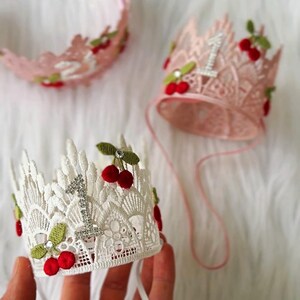 May include: Lace crowns with cherry accents, perfect for a first birthday celebration. The crowns are in pink and white, with a sparkling number "1" and red cherry details. One crown is on a headband.