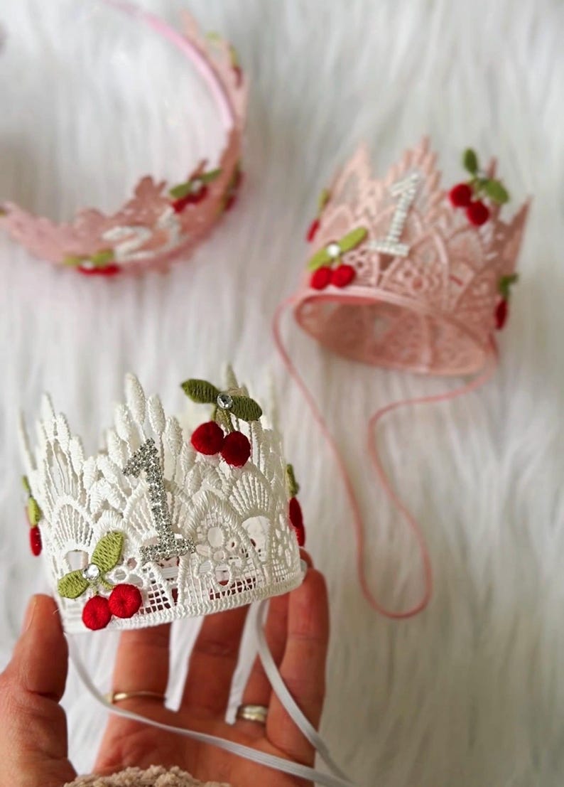 May include: Lace crowns with cherry accents, perfect for a first birthday celebration. The crowns are in pink and white, with a sparkling number "1" and red cherry details. One crown is on a headband.