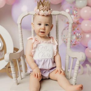 Baby girl wearing a gold lace first birthday crown with pink and lavender flowers and sparkly number one, seated in a pastel cake smash setup