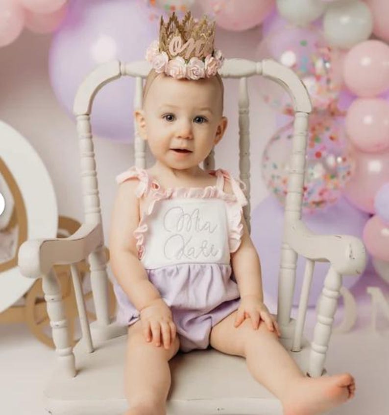 Baby girl wearing a gold lace first birthday crown with pink and lavender flowers and sparkly number one, seated in a pastel cake smash setup
