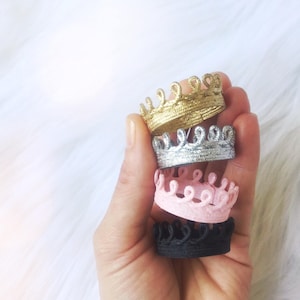 Hand holding a stack of four tiny lace crowns in metallic gold, silver, blush pink, and black. Each crown is mini sized, and made from sculpted lace. These crowns are part of a value bundle designed for pets, dolls, decor, or photography props. Perfect for kittens, hamsters, Barbie dolls, figurines, or party styling. Crowns do not have elastic or bands and are ideal for multipurpose use. Displayed against a soft white background.