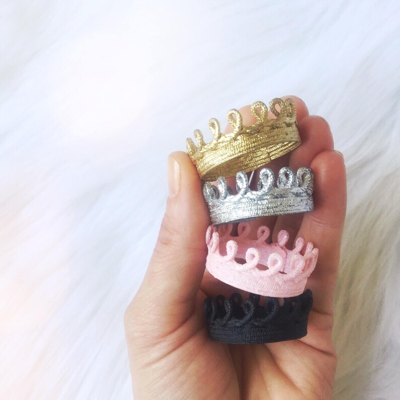 Hand holding a stack of four tiny lace crowns in metallic gold, silver, blush pink, and black. Each crown is mini sized, and made from sculpted lace. These crowns are part of a value bundle designed for pets, dolls, decor, or photography props. Perfect for kittens, hamsters, Barbie dolls, figurines, or party styling. Crowns do not have elastic or bands and are ideal for multipurpose use. Displayed against a soft white background.
