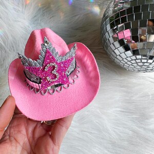 Mini pink cowboy hat birthday crown for baby girls and toddlers, featuring a sparkly silver tiara, hot pink glitter star, and rhinestone number 3 (customizable for any age). Ideal for cowgirl birthday outfits, pink pony parties, or My First Rodeo themes. Shown being held for scale with boho pampas grass in the background. Perfect for first birthdays, cake smashes, dress-up, or western-themed photo shoots.