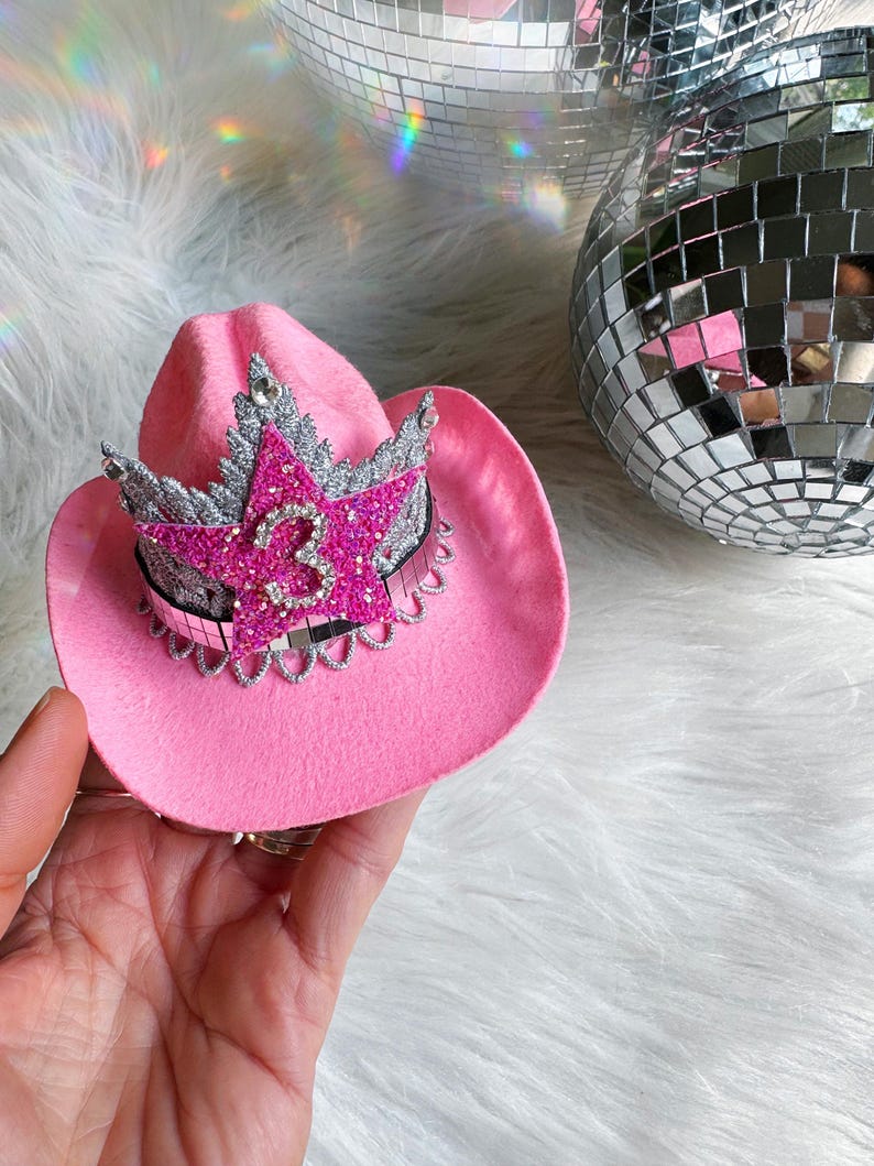 Mini pink cowboy hat birthday crown for baby girls and toddlers, featuring a sparkly silver tiara, hot pink glitter star, and rhinestone number 3 (customizable for any age). Ideal for cowgirl birthday outfits, pink pony parties, or My First Rodeo themes. Shown being held for scale with boho pampas grass in the background. Perfect for first birthdays, cake smashes, dress-up, or western-themed photo shoots.