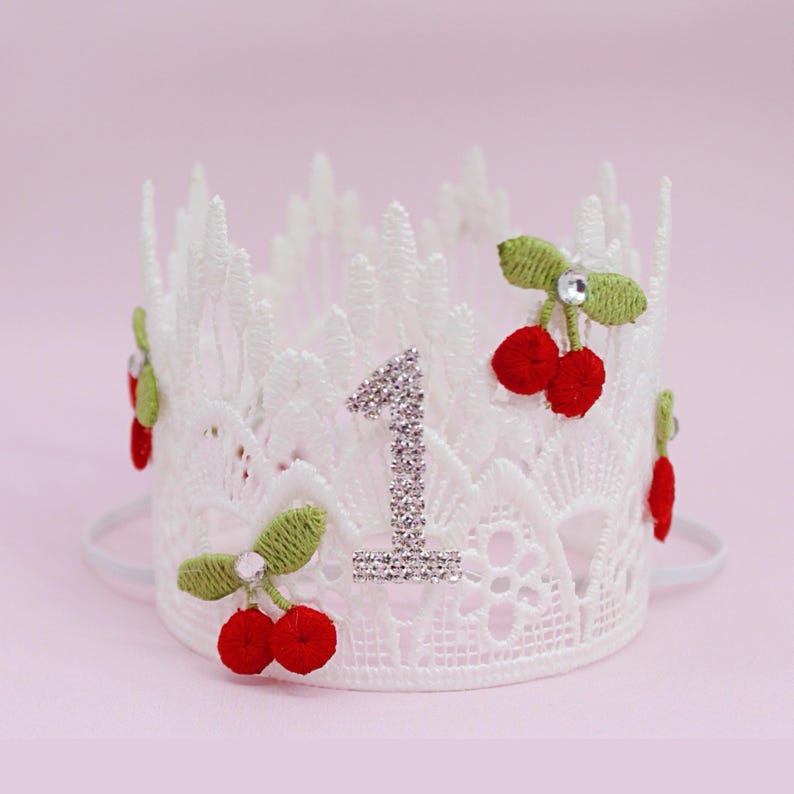 May include: White lace crown with a rhinestone number 1. The crown is decorated with red cherry accents and green leaves. A white elastic band secures the crown. Perfect for a first birthday celebration.
