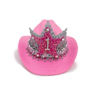 May include: A pink cowboy hat with a glittery star-shaped crown. The crown is embellished with silver accents and the number "1". The hat has a decorative silver trim around the brim, creating a festive accessory.