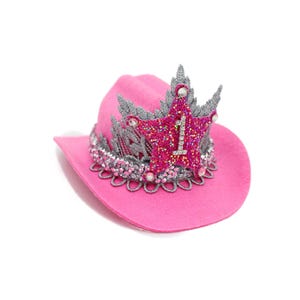May include: A bright pink cowboy hat with a decorative crown. The crown is silver and pink, with a star in the center that has the number "1" on it. The hat has a scalloped trim around the base.