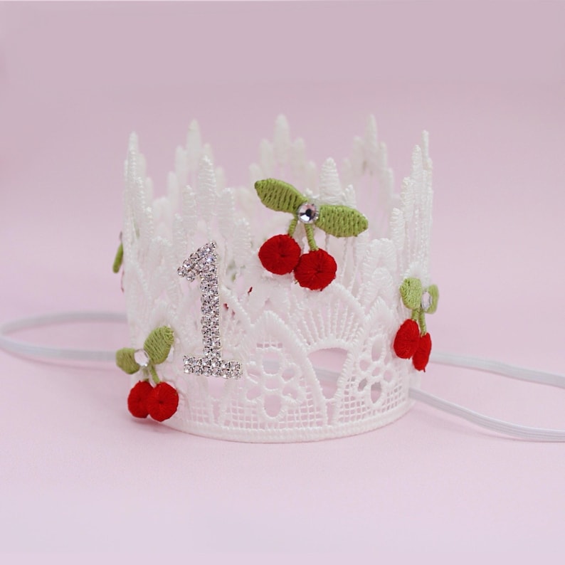 May include: White lace crown with the number "1" in silver rhinestones. The crown is decorated with red cherry accents and green leaves. A white elastic band is attached to the crown.