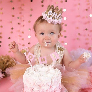 Smiling baby girl wearing a gold lace first birthday crown with pink floral accents, perfect for cake smash photos, milestone photography,  princess-themed birthday parties. A beautiful handcrafted 1st birthday tiara for an unforgettable celebration