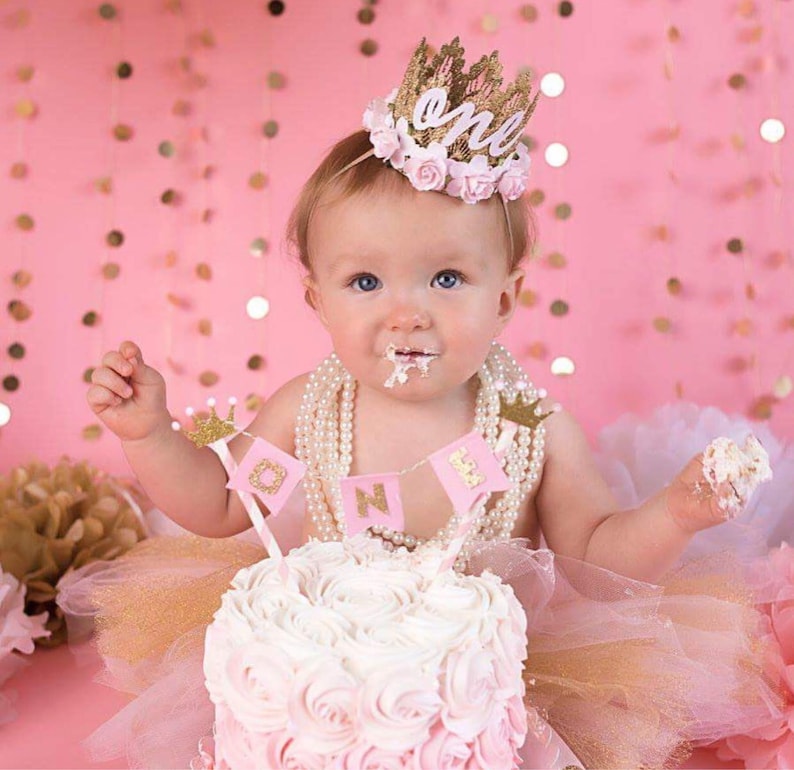 Smiling baby girl wearing a gold lace first birthday crown with pink floral accents, perfect for cake smash photos, milestone photography,  princess-themed birthday parties. A beautiful handcrafted 1st birthday tiara for an unforgettable celebration