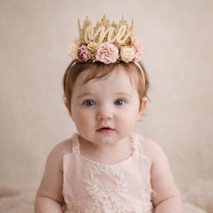 One year old baby girl wearing gold lace first birthday crown with cursive one script and blush pink and cream flowers, boho cake smash headband, soft neutral background