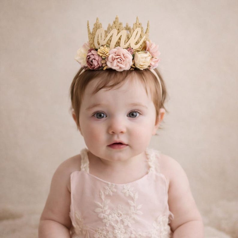 One year old baby girl wearing gold lace first birthday crown with cursive one script and blush pink and cream flowers, boho cake smash headband, soft neutral background