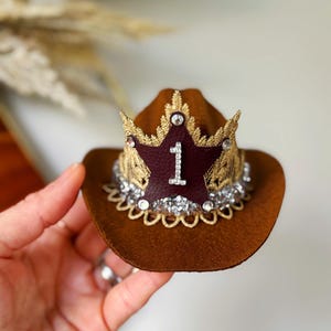 Mini brown cowboy hat made of soft felt with a gold faux leather star and metallic trim. The hat features a sparkly number one on the front for a first birthday theme. It’s lightweight, handcrafted, and designed for babies, toddlers, or small pets. Perfect for western birthday parties, rodeo photos, or dress-up costumes. Handmade in the USA by LoveCrush Crowns®.