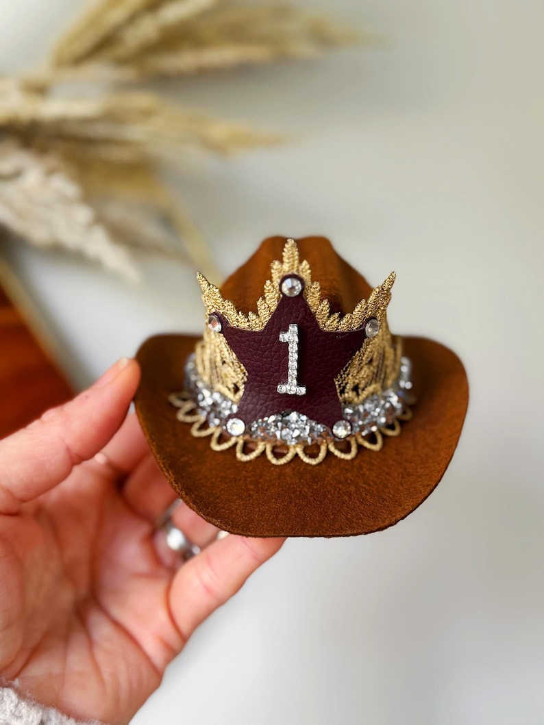 Mini brown cowboy hat made of soft felt with a gold faux leather star and metallic trim. The hat features a sparkly number one on the front for a first birthday theme. It’s lightweight, handcrafted, and designed for babies, toddlers, or small pets. Perfect for western birthday parties, rodeo photos, or dress-up costumes. Handmade in the USA by LoveCrush Crowns®.