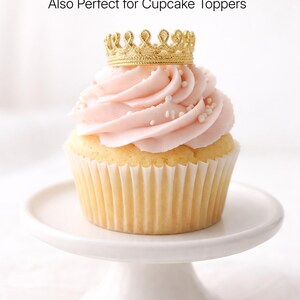 Mini gold lace crown cupcake topper on blush pink frosted cupcake with pearl sprinkles, displayed on white cake stand, tiny crown party decor, first birthday theme