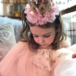 Baby girl wearing a gold lace first birthday crown with a blush pink tutu and soft floral accents. The perfect accessory for cake smash photography, ballerina-themed birthday parties, and princess dress-up. LoveCrush Crowns® designs one-of-a-kind heirloom-quality crowns for special memories.