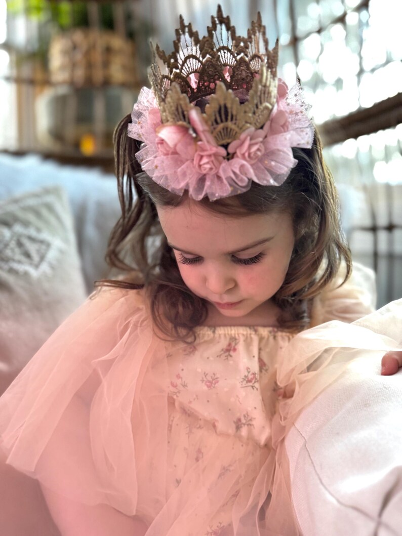 Baby girl wearing a gold lace first birthday crown with a blush pink tutu and soft floral accents. The perfect accessory for cake smash photography, ballerina-themed birthday parties, and princess dress-up. LoveCrush Crowns® designs one-of-a-kind heirloom-quality crowns for special memories.