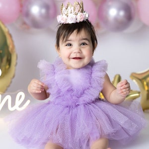 May include: A baby girl wearing a purple tulle dress with a sparkly finish and a crown that says "One" on it. The baby is smiling and looking at the camera.