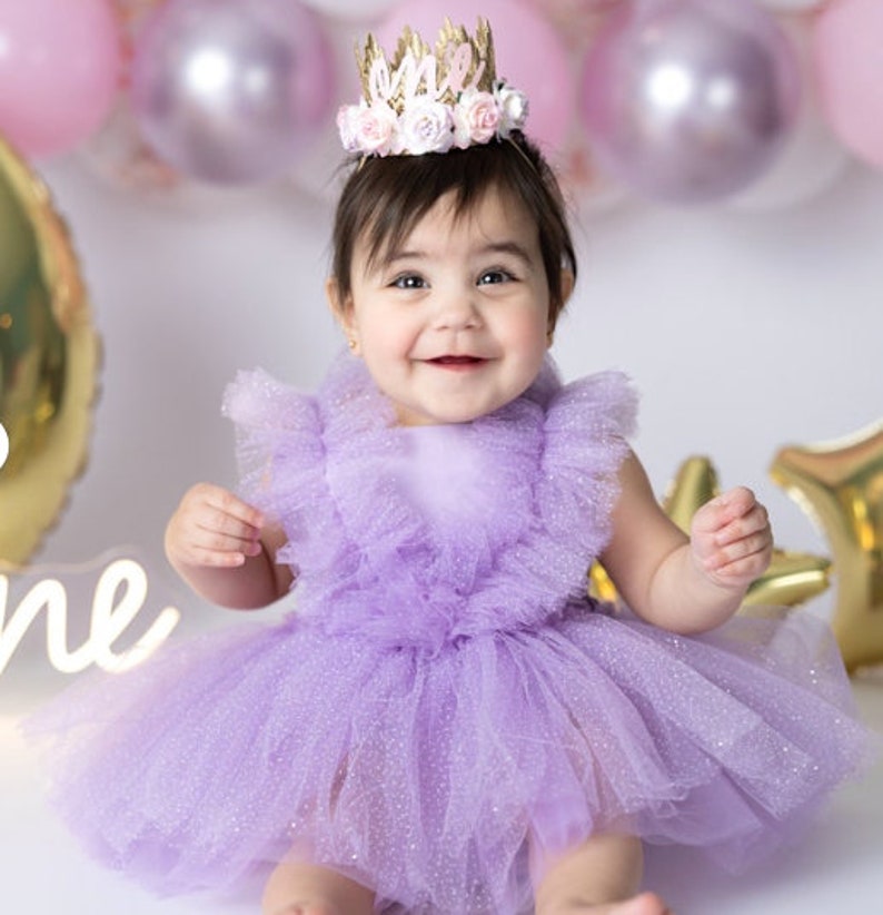 May include: A baby girl wearing a purple tulle dress with a sparkly finish and a crown that says "One" on it. The baby is smiling and looking at the camera.