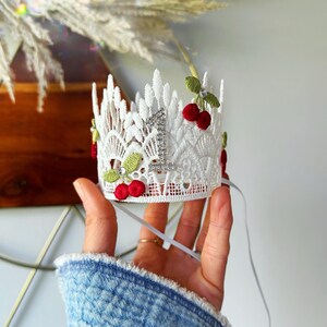 May include: A white lace crown with a silver number 1, adorned with red cherries and green leaves. The crown is held by a hand and has a ribbon attached. This festive crown is perfect for a first birthday celebration.