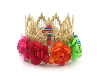 May include: A gold-colored crown with a floral design and the number "1" in the center. The crown is adorned with red, pink, orange, green, and blue flowers. The crown is likely for a first birthday celebration.