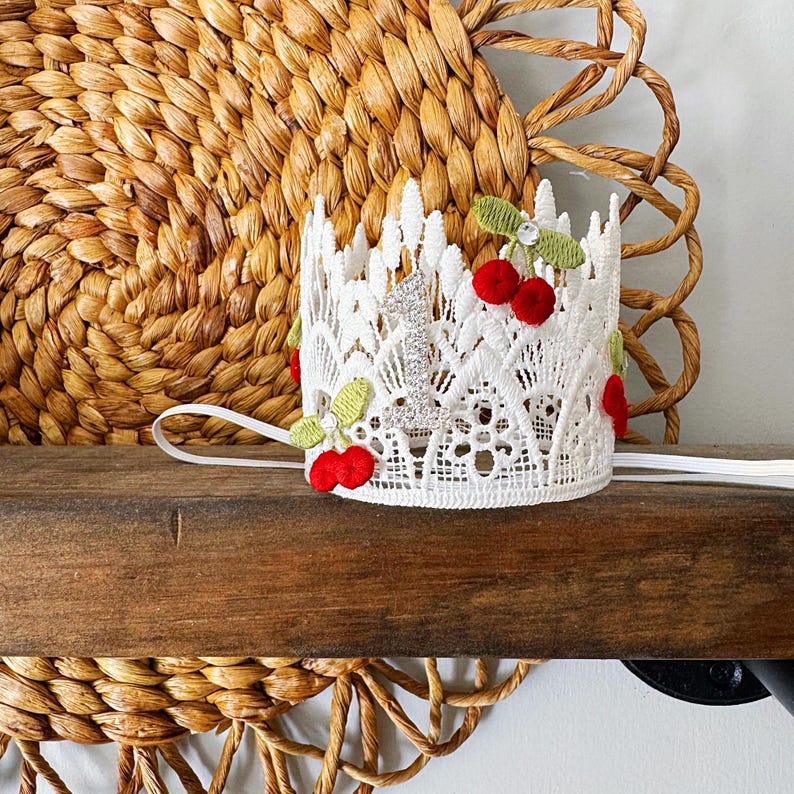 May include: A white lace crown decorated with red cherries and green leaves. The crown has a silver number "1" and a white elastic band. The crown is sitting on a wooden surface with a woven background.