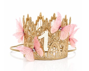 May include: A gold lace crown featuring a white glittery number one in the center. The crown is decorated with three soft pink butterflies with small clear crystals at their centers. A thin gold elastic band is attached to the base of the crown.