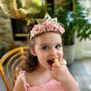 May include: A young girl wearing a pink dress with gold stars and a flower crown with a gold number two on top. She is eating a piece of cake.