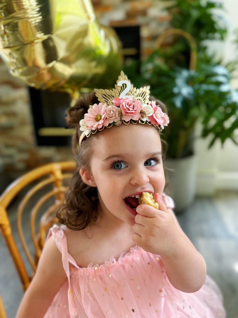 May include: A young girl wearing a pink dress with gold stars and a flower crown with a gold number two on top. She is eating a piece of cake.
