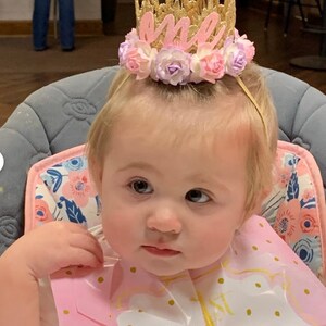 May include: A baby girl wearing a gold crown with pink flowers and the word "one" on it. She is wearing a pink bib with gold polka dots and is sitting in a high chair.