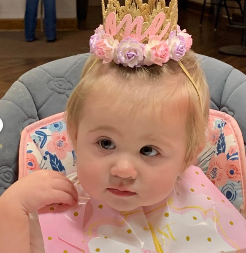 May include: A baby girl wearing a gold crown with pink flowers and the word "one" on it. She is wearing a pink bib with gold polka dots and is sitting in a high chair.