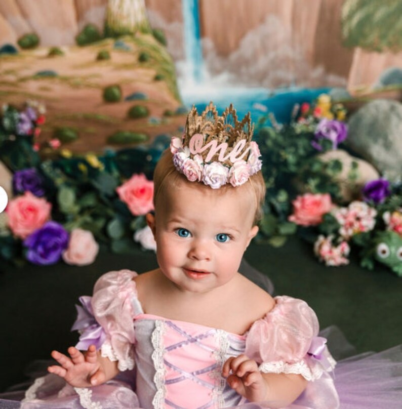 A young child wearing a pink and purple princess dress with a gold crown that says "one" on it. The child is surrounded by pink and purple flowers.