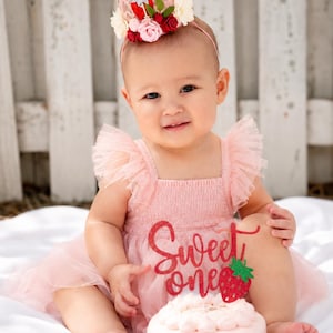 Baby girl wearing a Berry First Birthday Crown with strawberry floral details beside a pink cake for first birthday photos, berry sweet cake smash sessions, and strawberry-themed birthday celebrations.