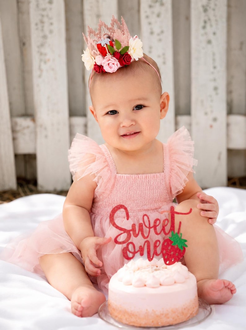 Baby girl wearing a Berry First Birthday Crown with strawberry floral details beside a pink cake for first birthday photos, berry sweet cake smash sessions, and strawberry-themed birthday celebrations.