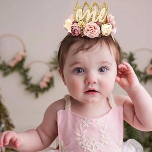May include: A baby girl wearing a pink dress with white floral embroidery and a gold crown with the word "one" and boho pink and white flowers.