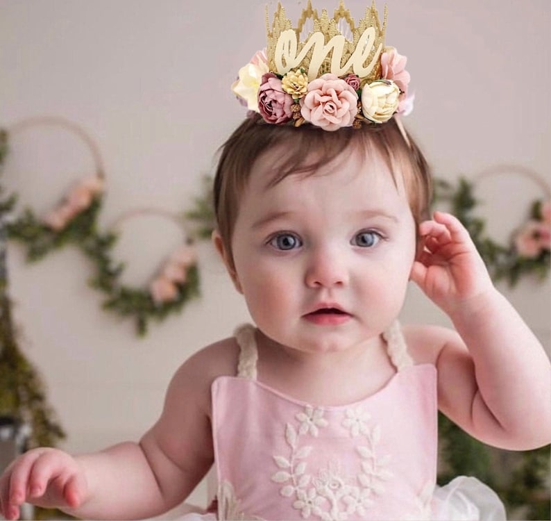 May include: A baby girl wearing a pink dress with white floral embroidery and a gold crown with the word "one" and boho pink and white flowers.