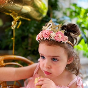 May include: A young girl wearing a pink dress with gold stars and a flower crown with the number "2" on it. She is eating a cookie and looking at the camera.