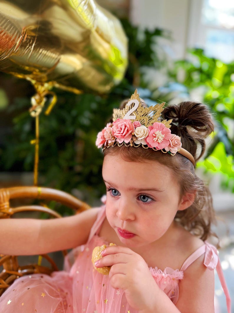 May include: A young girl wearing a pink dress with gold stars and a flower crown with the number "2" on it. She is eating a cookie and looking at the camera.