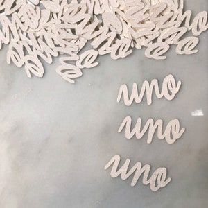 Puede incluir: Confetti blanco con purpurina con la palabra "uno" escrita tres veces sobre una superficie de m&aacute;rmol blanco.