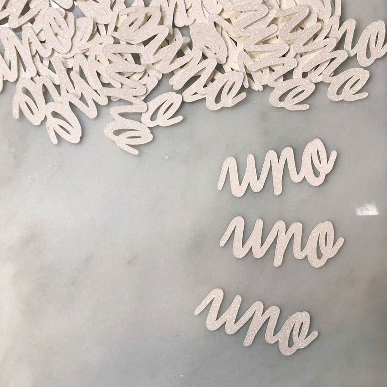 Puede incluir: Confetti blanco con purpurina con la palabra "uno" escrita tres veces sobre una superficie de m&aacute;rmol blanco.