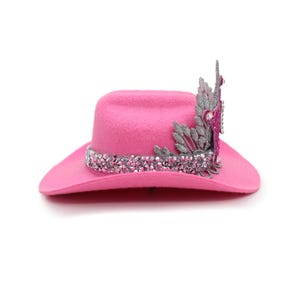 May include: A vibrant pink cowboy hat with a sparkling crystal band and decorative embellishments. The hat features a wide brim and a crown adorned with silver and pink accents. This stylish hat is perfect for adding a touch of flair to any outfit.