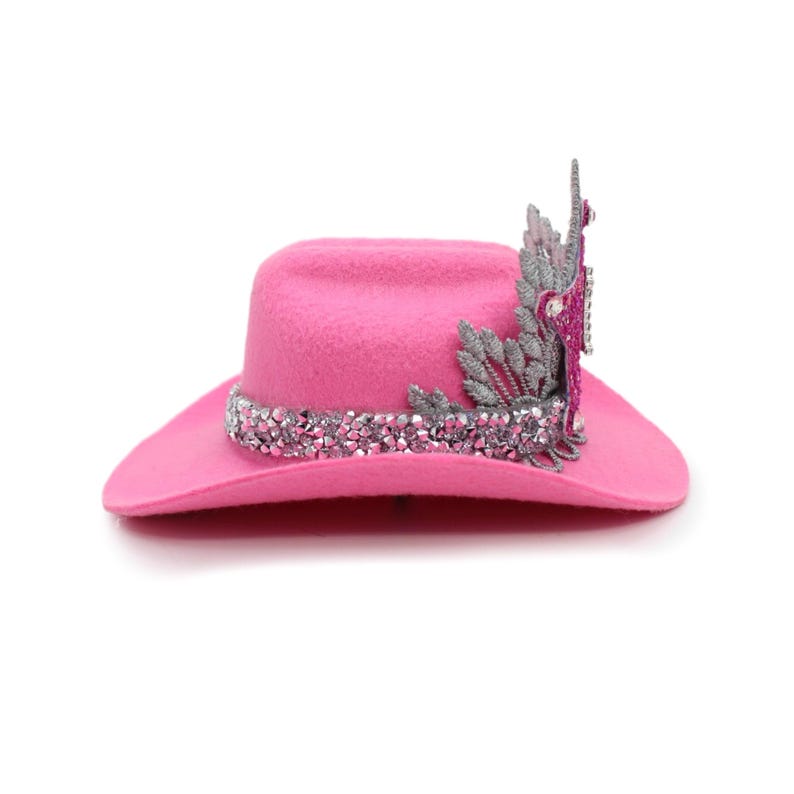 May include: A vibrant pink cowboy hat with a sparkling crystal band and decorative embellishments. The hat features a wide brim and a crown adorned with silver and pink accents. This stylish hat is perfect for adding a touch of flair to any outfit.