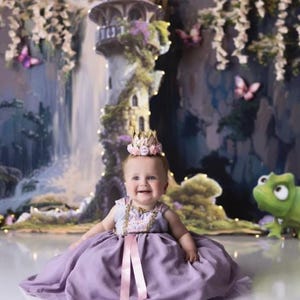 Smiling baby girl wearing a gold lace first birthday flower crown with pink and lavender roses, seated in a fairytale cake smash setup with pastel decor and whimsical backdrop