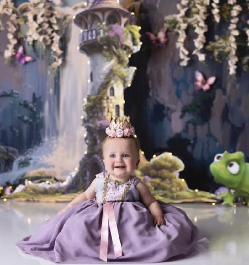 Smiling baby girl wearing a gold lace first birthday flower crown with pink and lavender roses, seated in a fairytale cake smash setup with pastel decor and whimsical backdrop