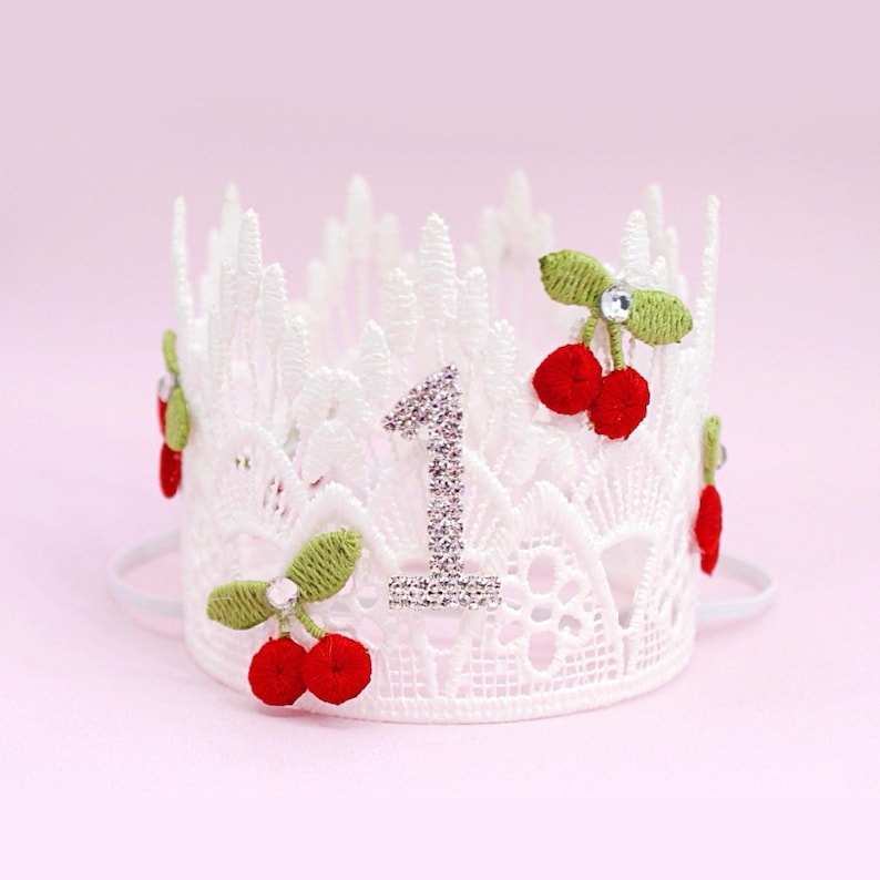 May include: White lace crown with red cherry accents and a rhinestone number 1. The crown has a delicate design with a white elastic band. The cherries have green leaves and a small rhinestone.