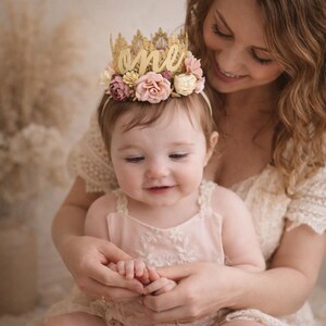 One year old baby girl wearing gold lace first birthday crown with cursive one script and blush pink floral details, sitting with mother, boho cake smash photo, soft neutral background