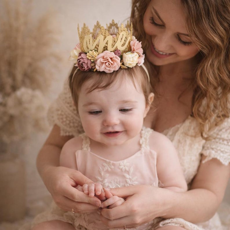 One year old baby girl wearing gold lace first birthday crown with cursive one script and blush pink floral details, sitting with mother, boho cake smash photo, soft neutral background