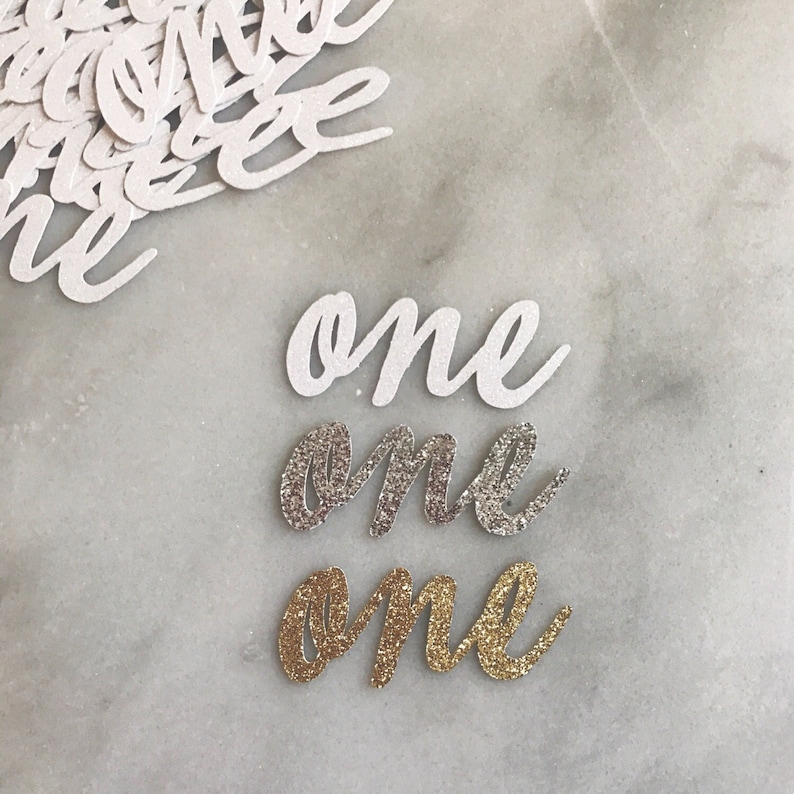 Puede incluir: Tres recortes de papel de la palabra "one" en diferentes estilos. El recorte superior es blanco, el recorte del medio es de purpurina plateada y el recorte inferior es de purpurina dorada.